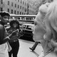 On the left side of frame, three Black women stand together on the street; on the right, a blond woman walks by with a serious face, and a man's leg is in the background