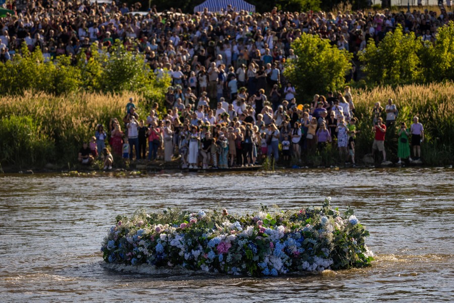 A large crowd stands on the banks of the a river, watching as a huge floral wreath floats past.