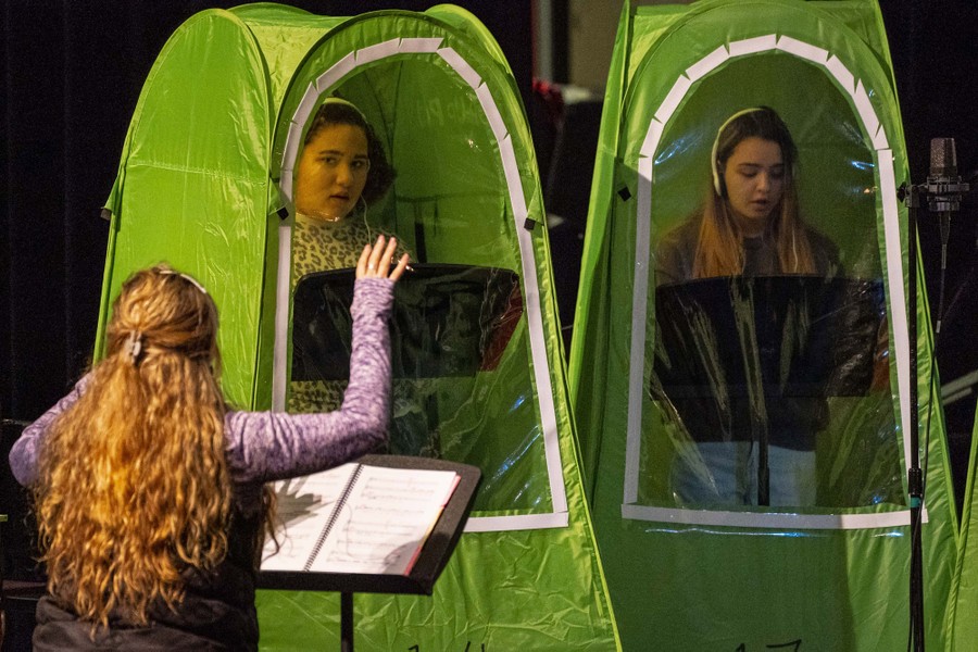 Two students sing in a music class, standing inside tall individual-sized tents with clear plastic windows.