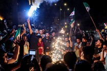 Syrians in a crowd light sparklers and wave flags on the street.