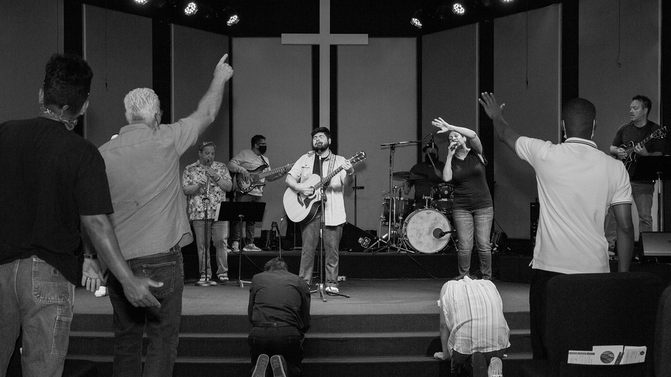 A black-and-white photo of a group standing in a church, listening to someone at the front playing a guitar