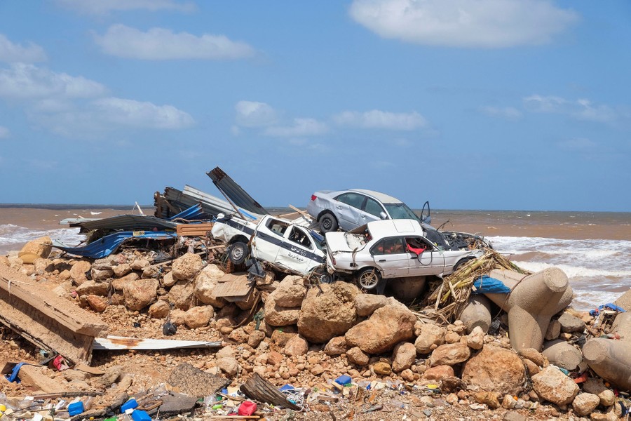 Several damaged cars rest on a large pile of rocks, among other debris, along a coastline.