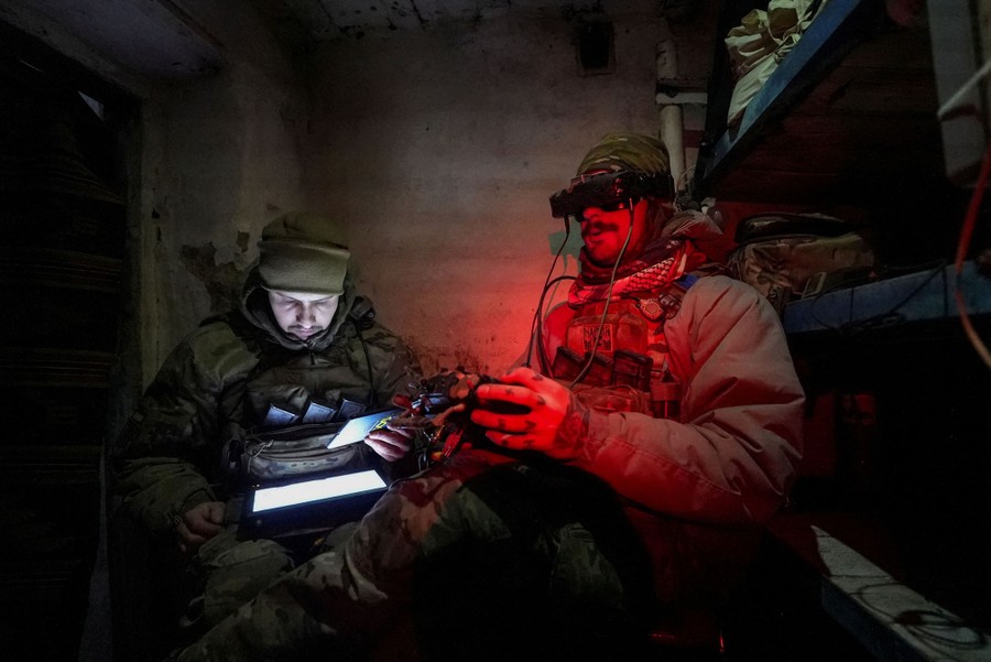 Two soldiers sit in a concrete room, using a tablet, VR goggles, and a controller to pilot a remote drone.