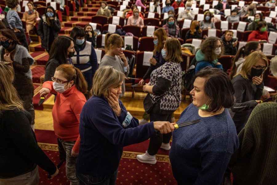 Dozens of women watch from their seats in an auditorium as more than a dozen other women practice defending themselves with replica knives.
