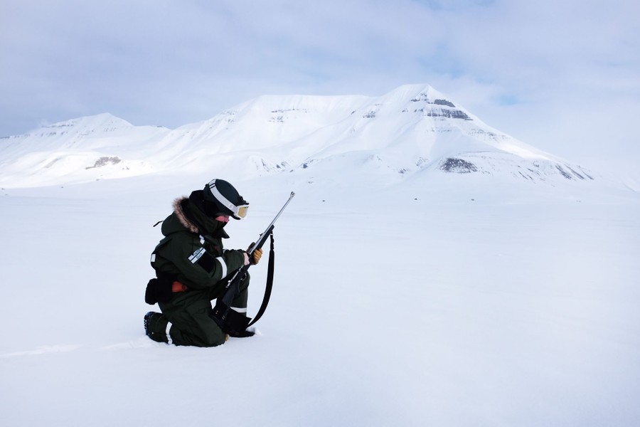 A person kneels to load a rifle in a snowy, treeless landscape.