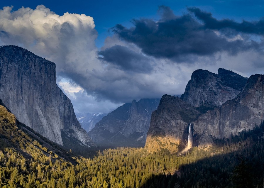 Sunlight illuminates pine trees across the floor of Yosemite Valley, surrounded by steep cliffs, mountains, and a tall waterfall.
