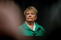 A photograph of Janet Protasiewicz, a short-haired blond woman, in a green jacket standing in front of a microphone