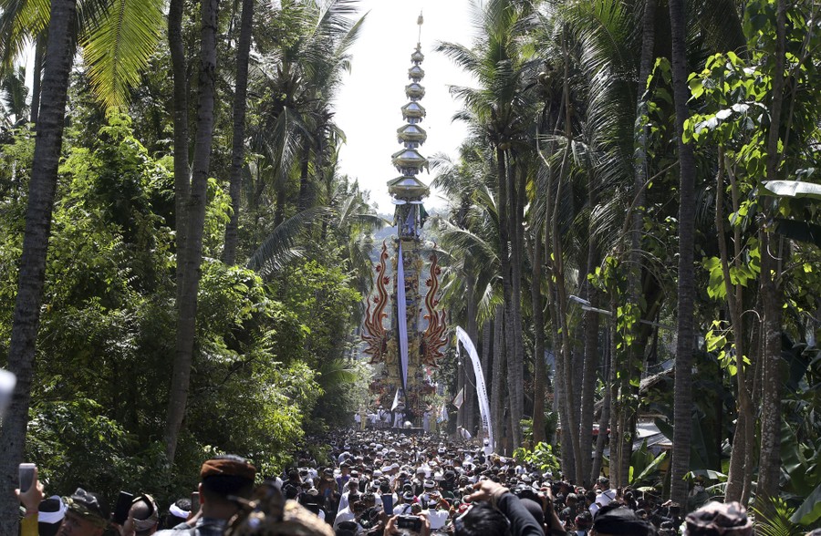A crowd of people parades behind a very tall ornamental tower, among palm trees.