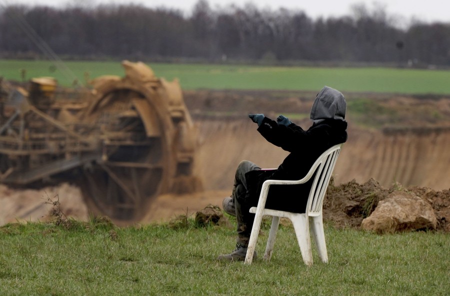 A person sits in a white-plastic chair in a field at the edge of an open coal-mine pit, with a large excavator visible in the background.