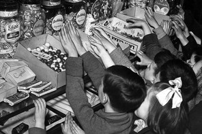 A black and white photograph of a gaggle of children reaching up onto a counter filled with boxes and jars of candy