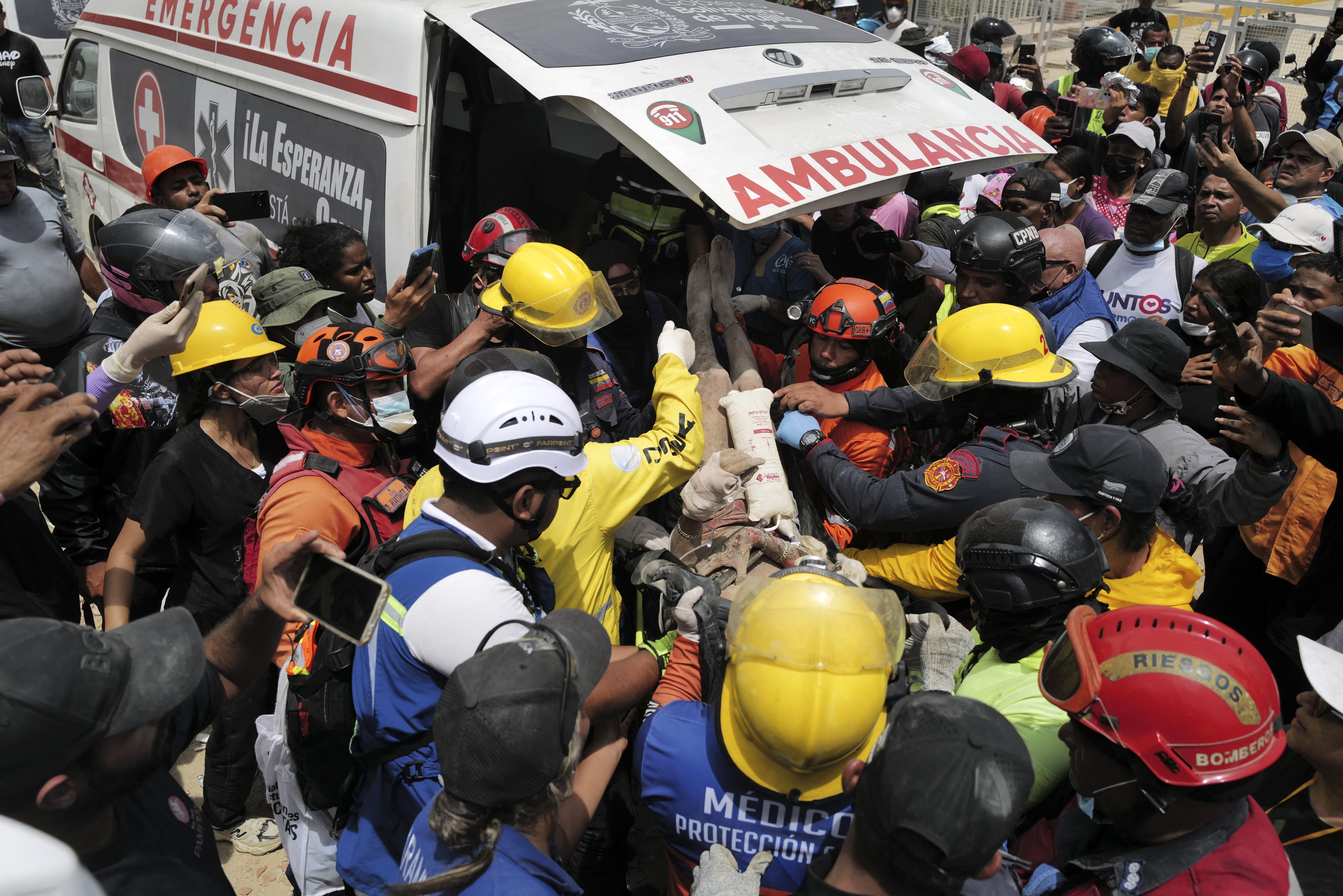 Rescuers, volunteers, and onlookers crowd around an ambulance as a survivor is removed from earthquake rubble.