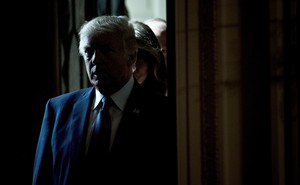 A 2017 photo of then-President Donald Trump wearing a blue suit and tie, standing in a dark corridor