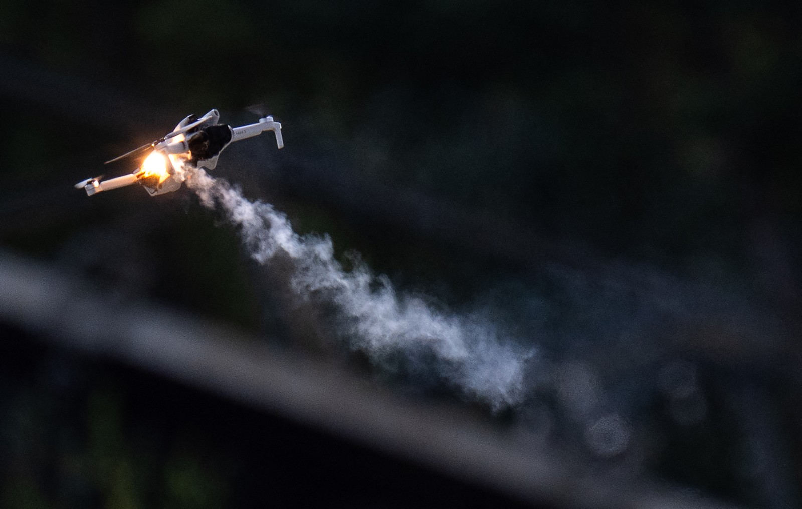 A small quad-rotor drone flies overhead, smoke trailing from its body, after being hit by a laser weapon.