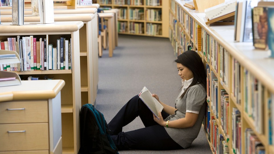 Asian American student sitting on the floor of a library reading a book, leaning back against the shelves