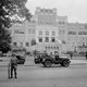 This black and white photo shows men in combat attire standing guard outside a large brick school building. Three military-esque Jeeps are in the photo.