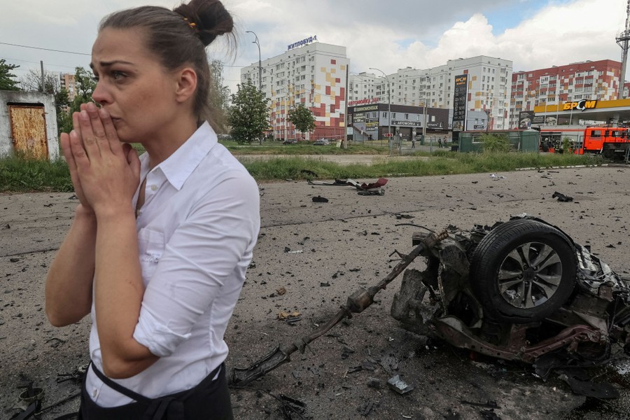 A civilian stands, hands to their face, beside the scattered wreckage of a bomb-damaged vehicle.