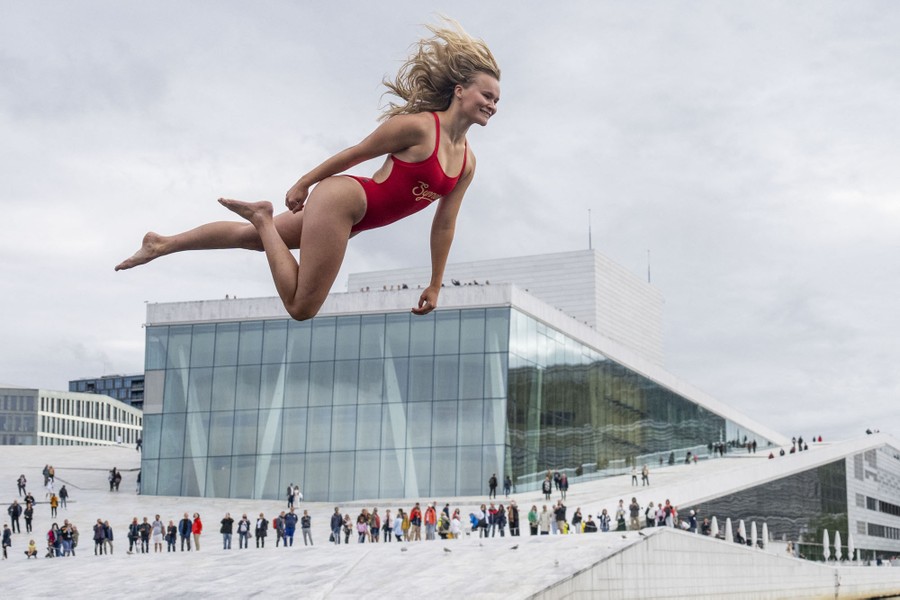 A person in a swimsuit in mid-air, practicing a high dive in front of a modern opera house