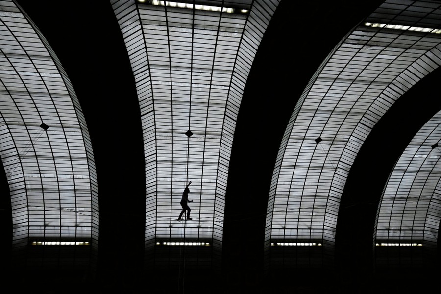 A person walks a tightrope inside a large space, beneath an arched roof.