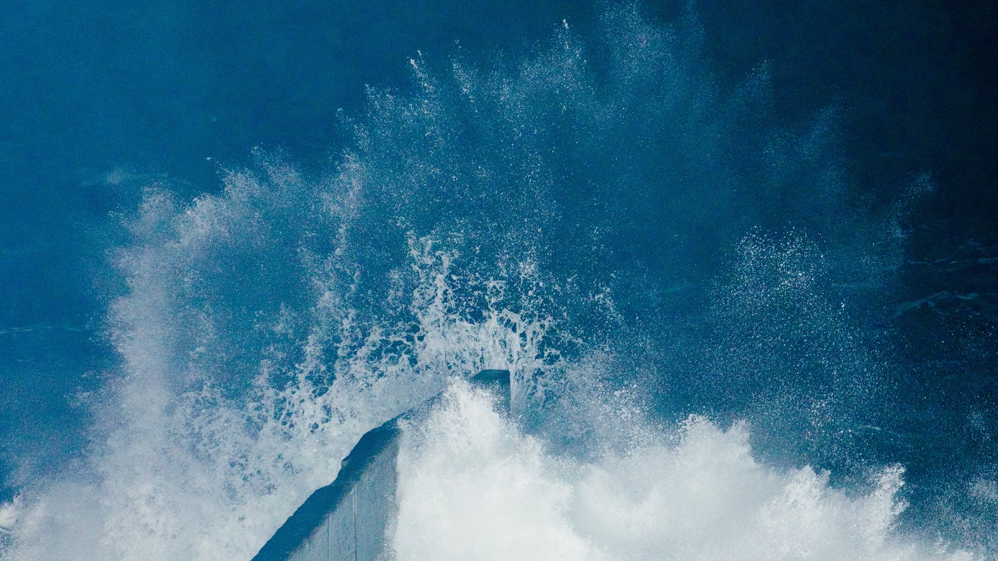 A color photograph of a wave breaking against a concrete barrier