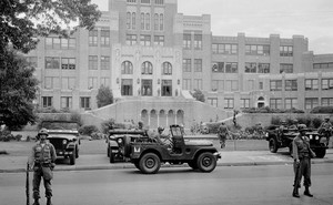 This black and white photo shows men in combat attire standing guard outside a large brick school building. Three military-esque Jeeps are in the photo.