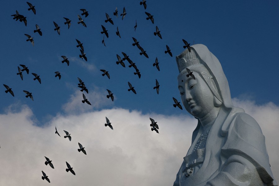 Pigeons fly in front of a very large statue of a Buddhist deity.