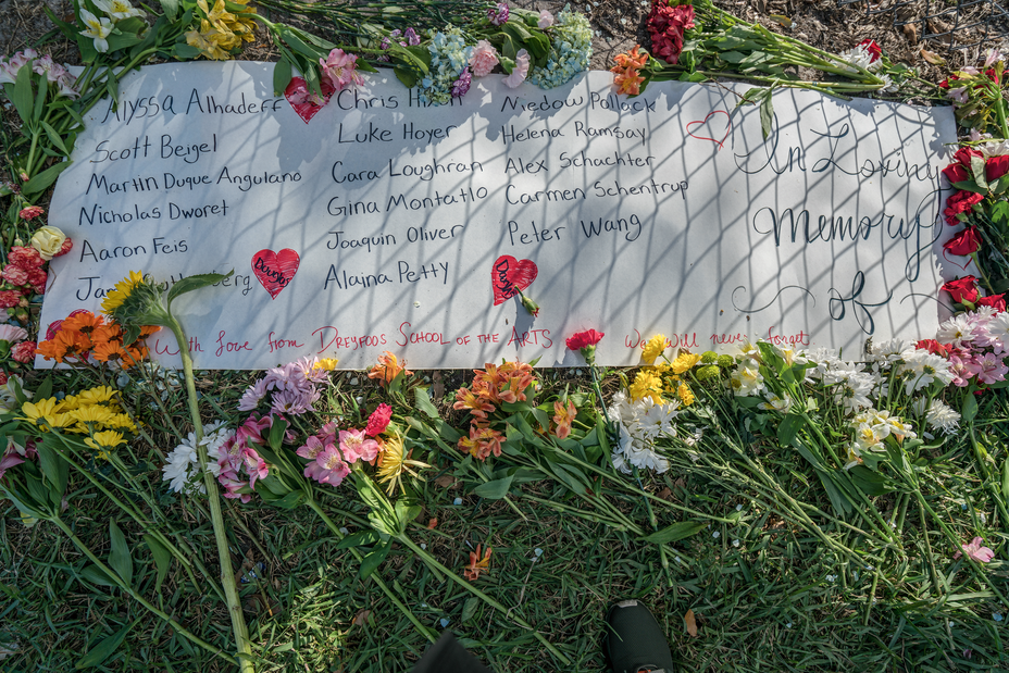 photo of a hand-drawn banner lying on grass outside that included "In Loving Memory of," a list of student names, and red hearts, surrounded by dozens of loose flowers