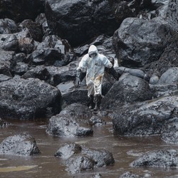 A worker wearing protective gear walks on oil-covered rocks beside spilled oil in a cove.