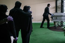 A funeral procession in New Jersey. A black man wearing a mask is pushing a casket.