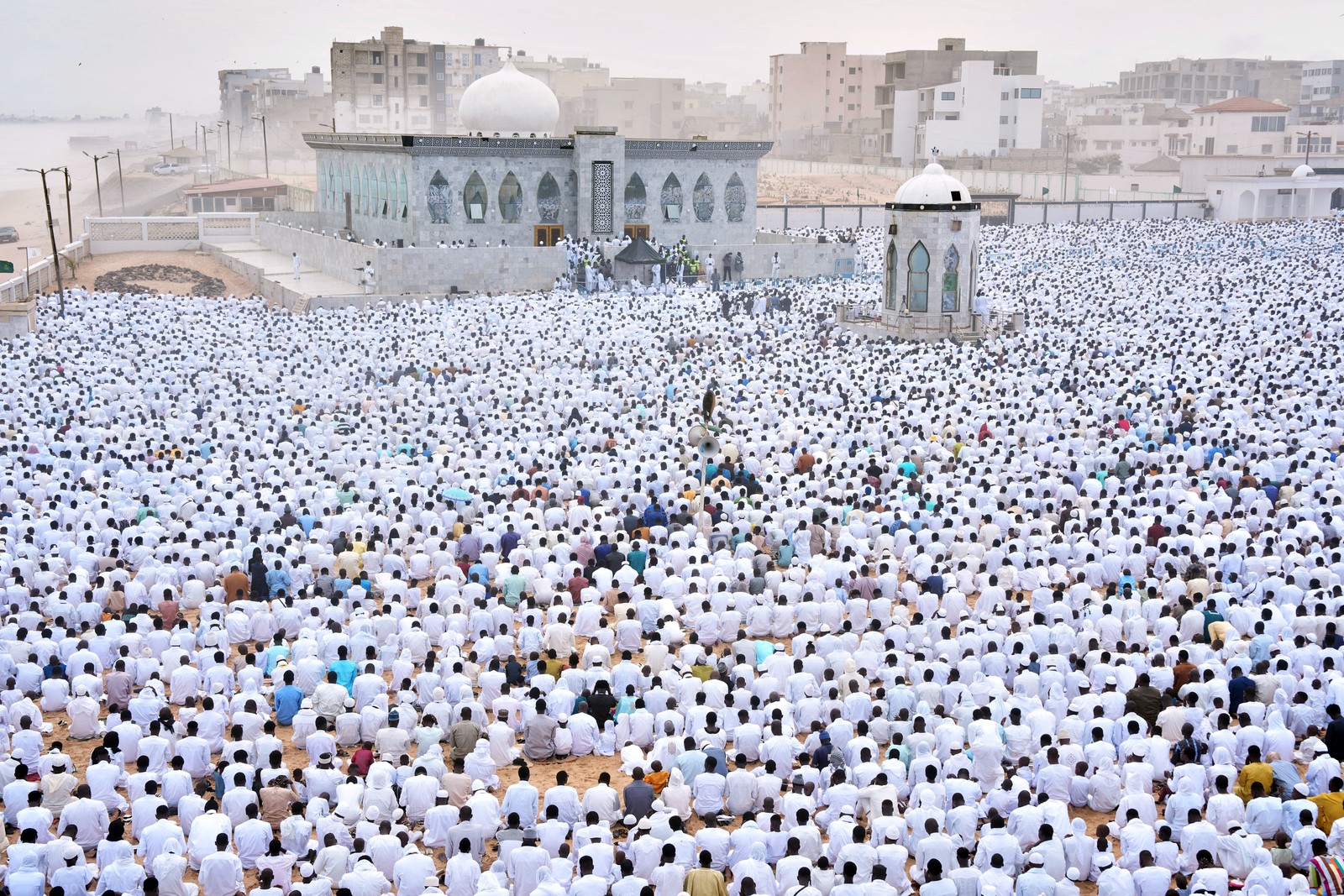 A large crowd of people, mostly dressed in white, sits in rows in front of a mosque.