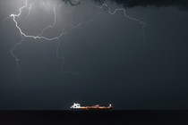 Color photograph of a huge lightning formation lighting up the night sky above an oil tanker