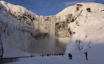 A couple dozen tourists in winter gear stand around beneath a tall cliff and waterfall, surrounded by snow and ice.