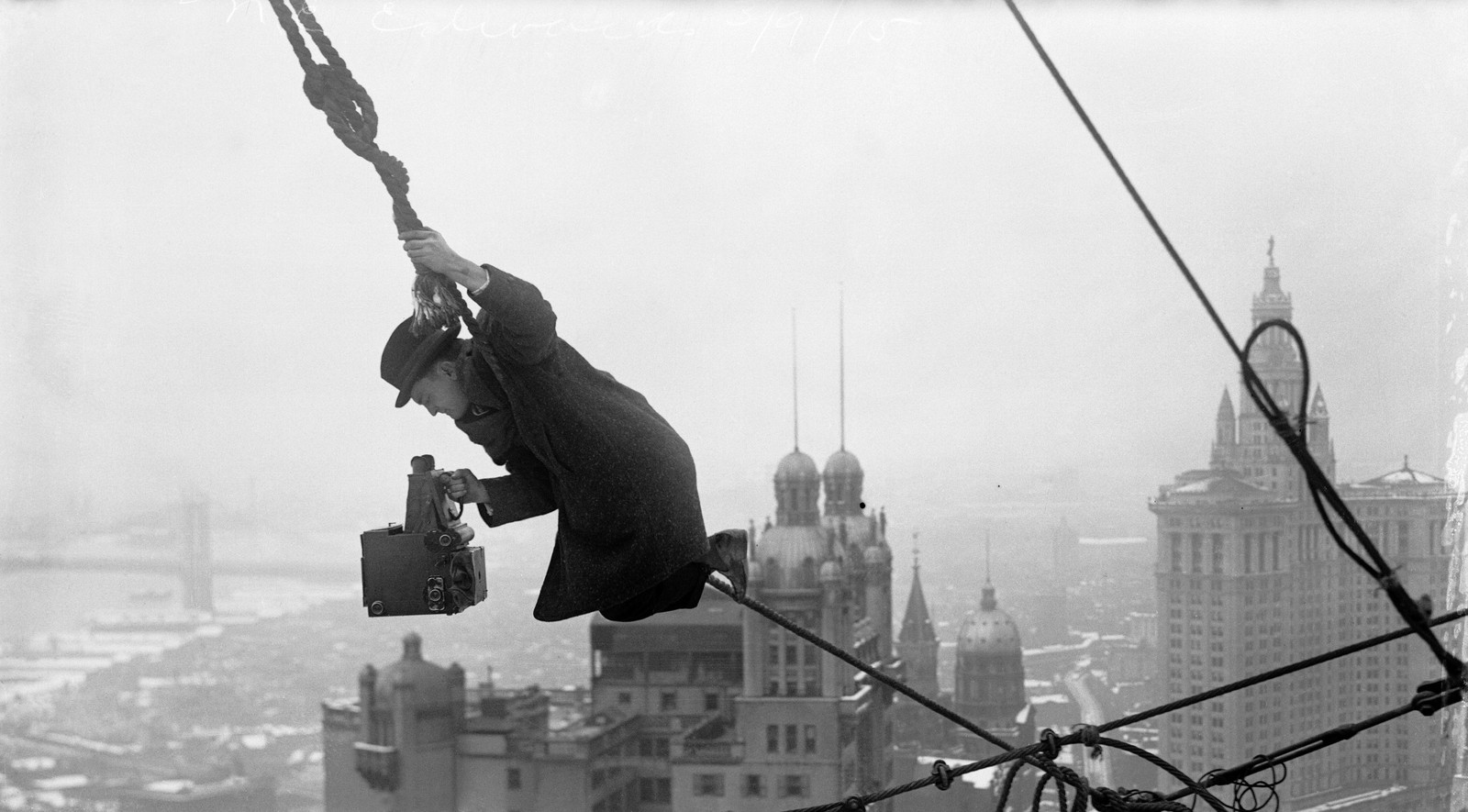 A photographer holding a large boxy camera hangs perilously from a rope on a high building, with a cityscape visible in the background.