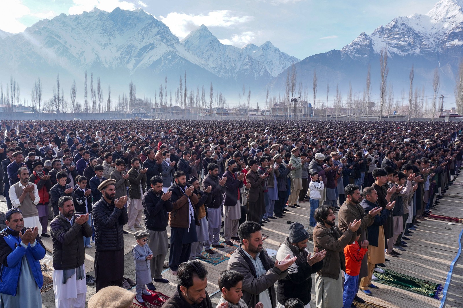 A large crowd of Muslims pray together, standing outside, with tall, snow-covered mountains in the background.