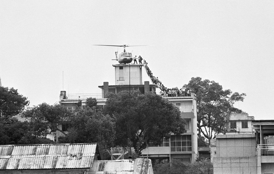 A line of people climb a steep staircase on a building's roof to board a helicopter that has landed on a very small rooftop space.