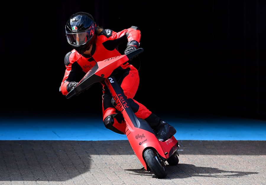 A person wearing a helmet and racing suit leans into a turn while riding an electric scooter.