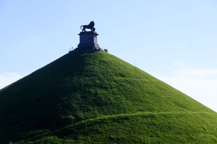 A statue of a lion stands on a plinth atop a tall conical hill covered in grass.