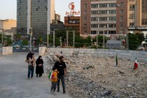 A Iranian family walks by a field of rubble in an urban neighborhood, the young boy carrying an Iranian flag.