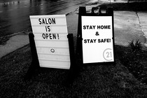 A photo of two road-side signs. One says, "Salon is open!" The other says, "Stay home and stay safe!"