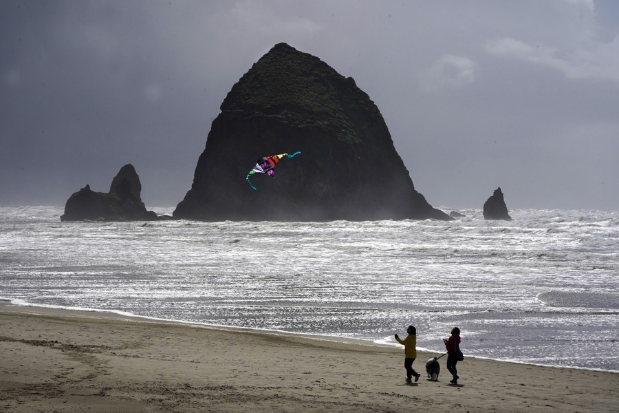 Two people fly a kite on a beach near a tall rock formation.