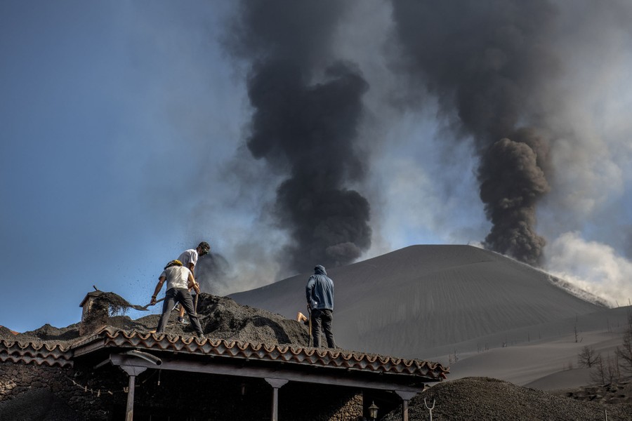 Several people use shovels to clear piles of ash off the roof of a house near an erupting volcano.