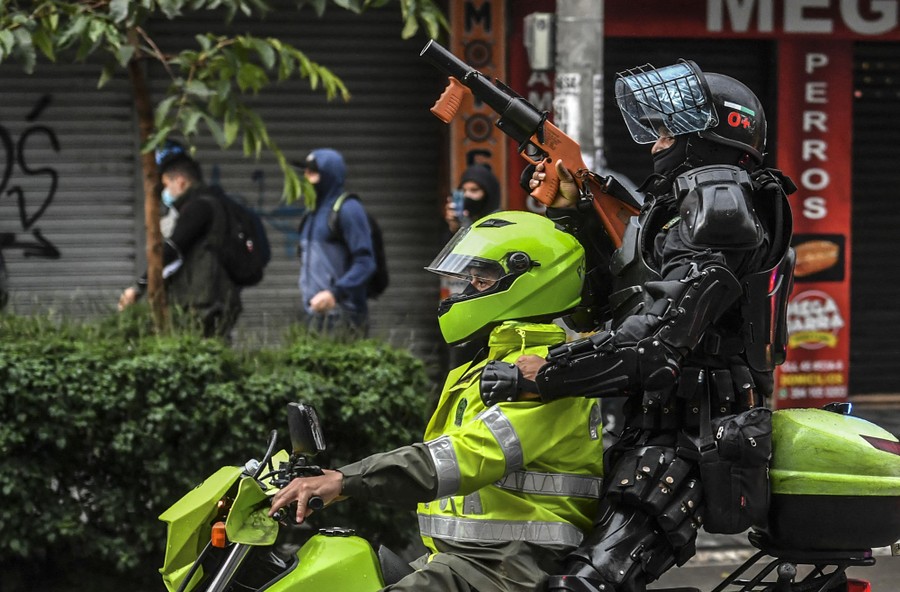 Two riot police officers in uniform ride a motorcycle. One of them holds up a crowd-control weapon.