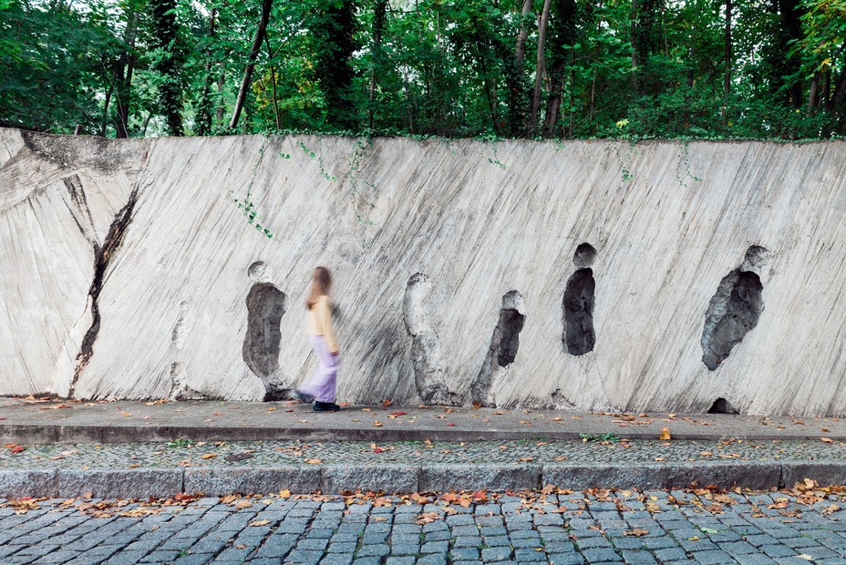 photo of person walking past wall with abstract human figures etched in it, with trees and street
