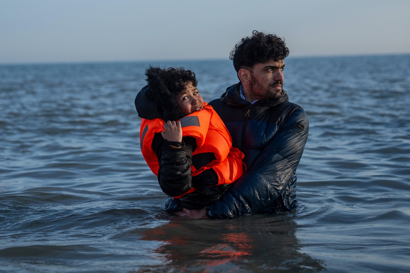 A boy wearing a life preserver is carried by a man through waist-deep water.