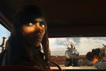 A film still showing Anya Taylor-Joy as the character Furiosa looking backward while sitting in a car; other cars, and an explosion, can be seen through the windshield in the desert landscape behind her.