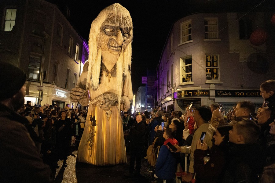 A very tall puppet of a wild-looking woman is guided through a crowd during a parade.