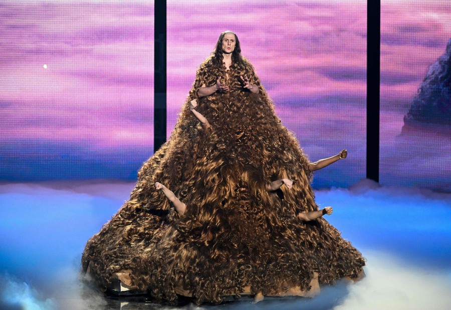 A person in a long and hairy costume performs. At least seven human arms are emerging from the costume.