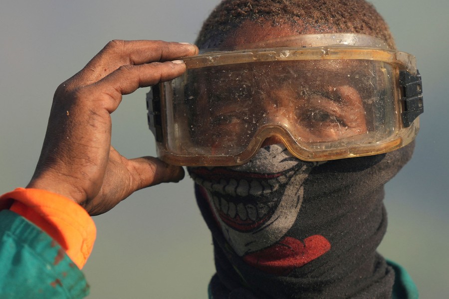 A man wearing a face mask with a toothy grin on it adjusts his protective goggles amid thick smoke from a fire.