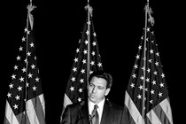 Black-and-white photo of Ron DeSantis speaking into a microphone, in front of three American flags