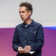 Malcolm Gladwell stands in front of a purple-and-blue screen giving a speech.