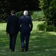 Former Presidents Barack Obama and Bill Clinton walk through a garden, facing away from the camera.